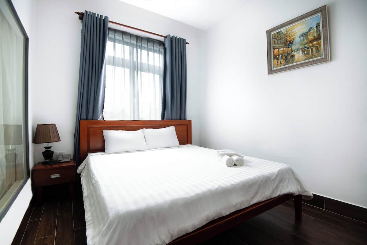 Hotellihuone, jossa on puinen sänky, valkoiset vuodevaatteet, yöpöytä lampun kanssa ja taulu seinällä.
