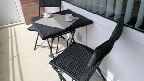 A small outdoor balcony with a black metal table and two chairs, a white cup on the table, and a view of a car through the window. - Sylter Leuchtfeuer (Sylt)