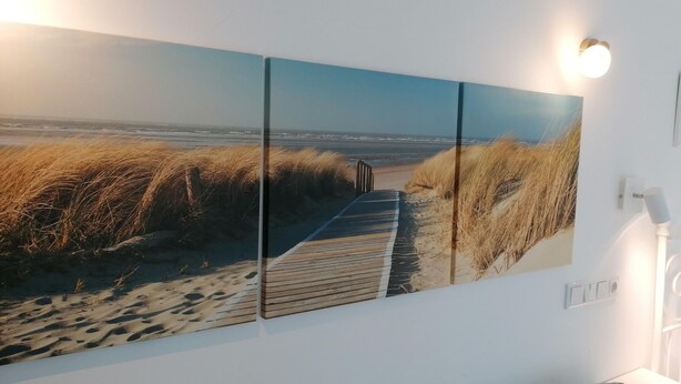 A corridor with a large, framed beach scene on the wall, a white metal bed frame, and a wall-mounted light fixture. - App. Friesenkoje (Sylt)