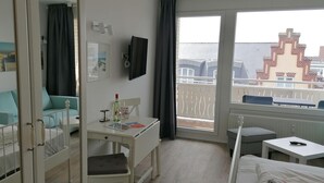 A hotel room with a bed, a desk, a chair, a TV, and a balcony with a view of buildings. - App. Friesenkoje (Sylt)
