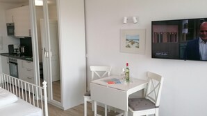 A modern hotel room with a dining area, a TV mounted on the wall, and a kitchen visible through an open door. - App. Friesenkoje (Sylt)