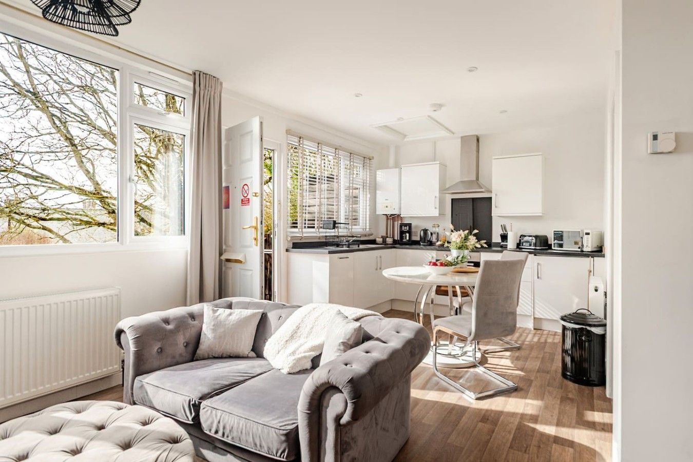 A modern kitchen with white cabinets, a dining table, and a grey sofa in a bright room with large windows.