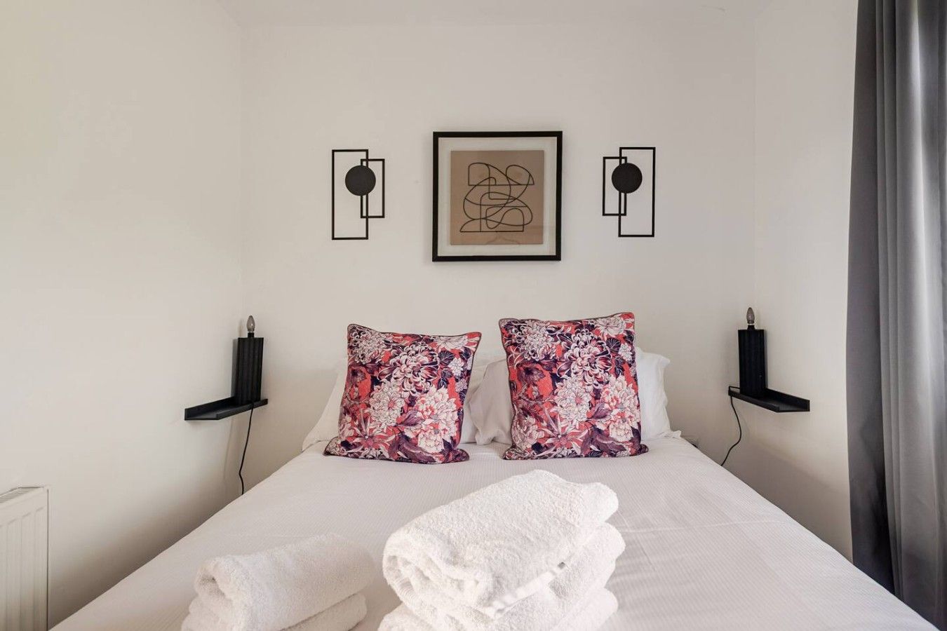 A hotel room with a bed, two pillows, a framed artwork, and a wall-mounted shelf.