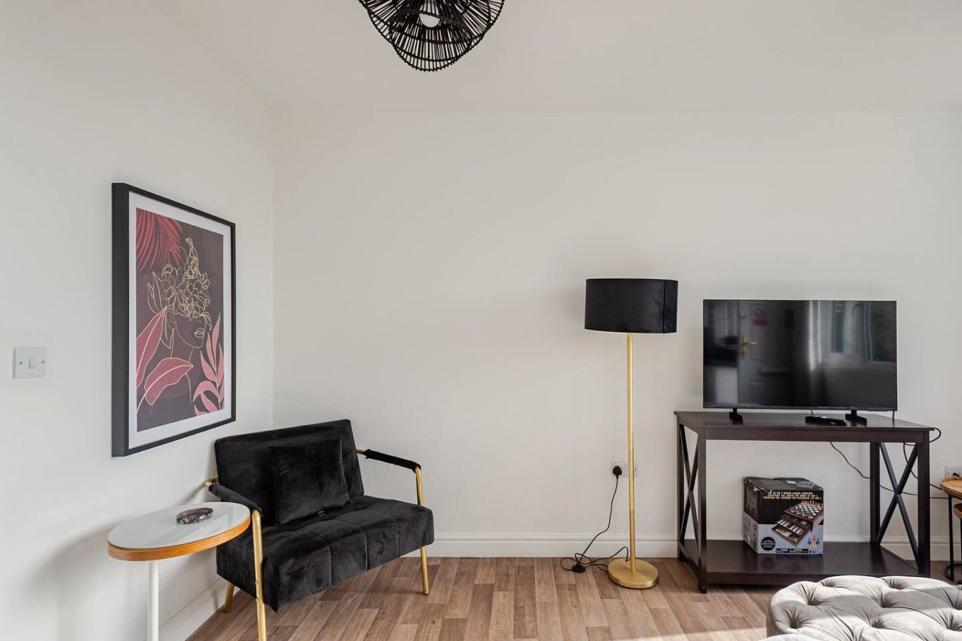 Un salon avec un canapé noir, une petite table ronde, un lampadaire, un téléviseur posé sur un meuble et un tableau encadré au mur.