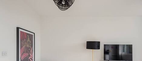 Ein Wohnzimmer mit einem schwarzen Sofa, einem kleinen runden Tisch, einer Stehlampe, einem Fernseher auf einem Schrank und einem gerahmten Bild an der Wand.