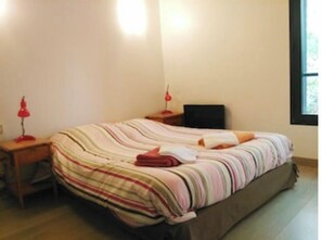 Ein Schlafzimmer mit einem Bett, einem Nachttisch, einem Schreibtisch, einem Sessel, einer Lampe und einem Fenster.