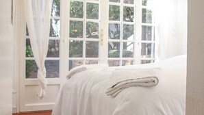 Een slaapkamer met een groot raam, een bed met witte lakens en een gordijn.