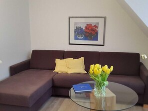 Uma sala de estar com um sofá marrom, uma almofada amarela, uma mesa de centro de vidro, um vaso com flores amarelas e um quadro na parede.