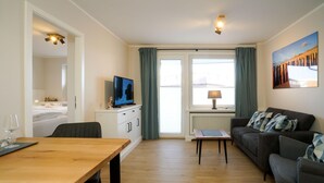 A modern living room with a wooden dining table, a sofa, a TV, and a window with curtains. - Sylter Krebs, Haus Ankerlicht, Wohnung 65 (Sylt)