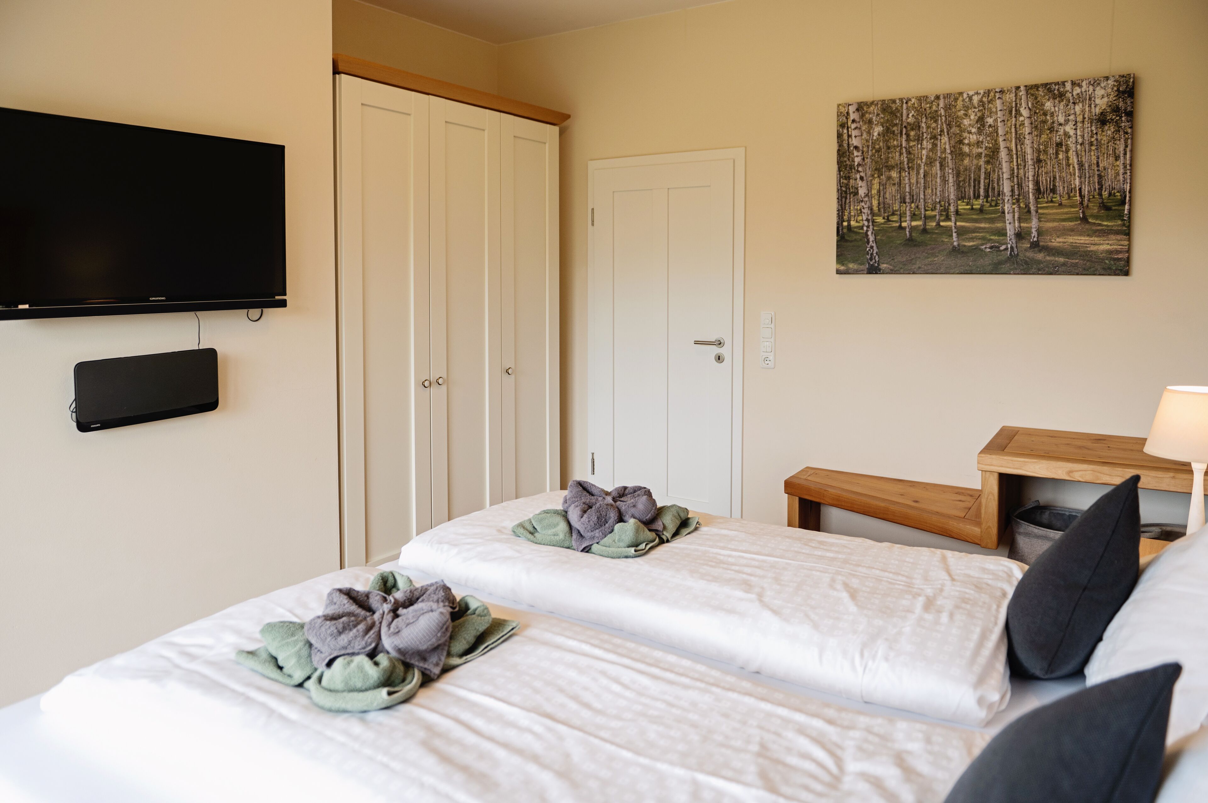 A hotel room with two beds, a TV, a wardrobe, and a painting of a forest.