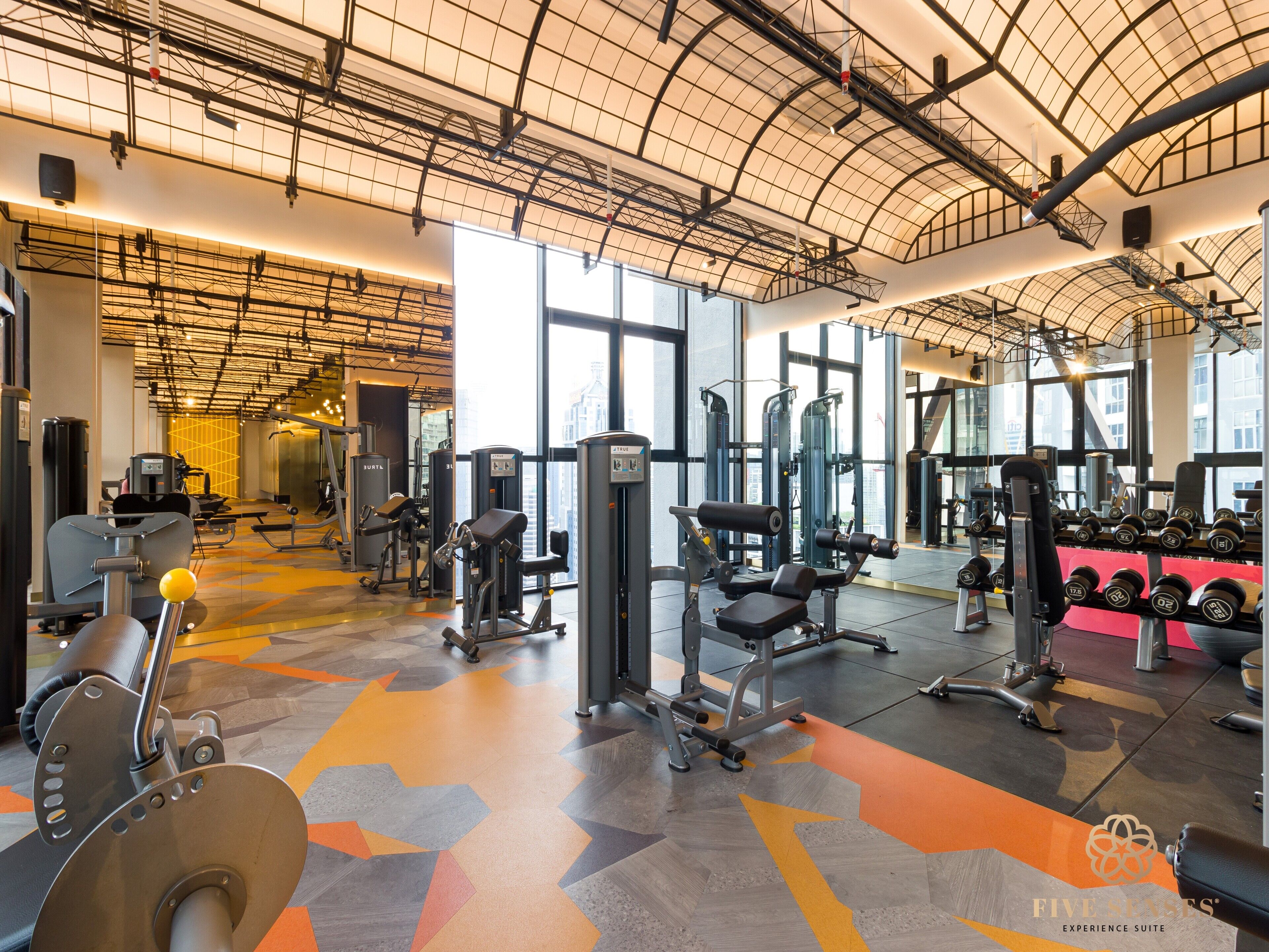 A modern gym with various exercise equipment, large windows, and a high ceiling.