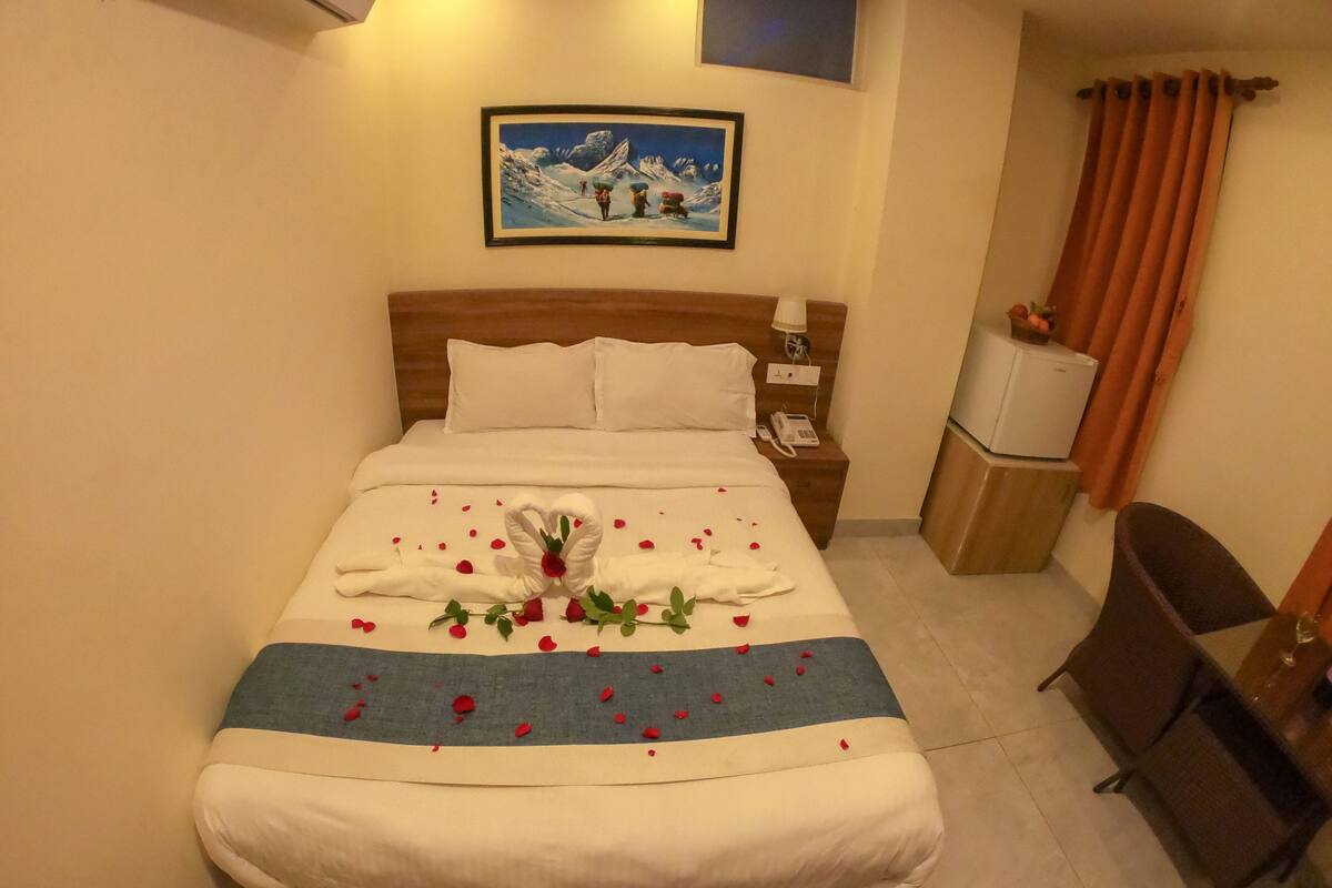 A neatly made bed with a heart-shaped towel, rose petals, and a small bouquet. There is a painting on the wall above the bed and a television on a wooden cabinet to the right.