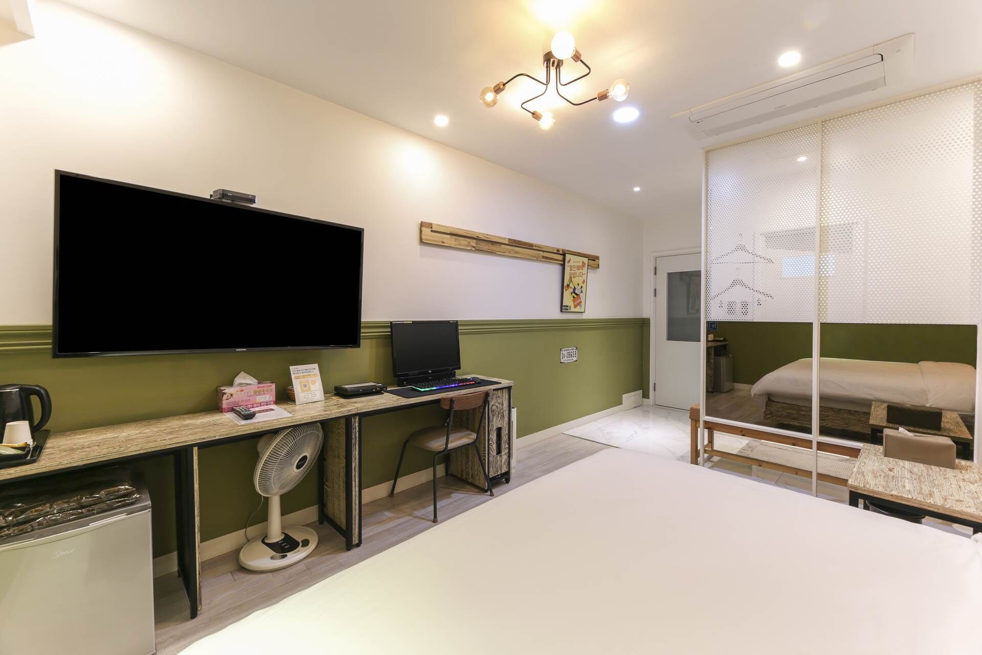 Habitación de hotel con televisión de pantalla plana, escritorio con una computadora portátil, refrigerador pequeño, cama con colchón blanco y ventana con persianas.