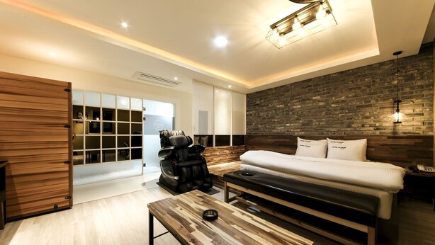 A modern hotel room with a wooden bench, a massage chair, a bed with white linens, and a brick accent wall. - The Stay Series (Daegu)