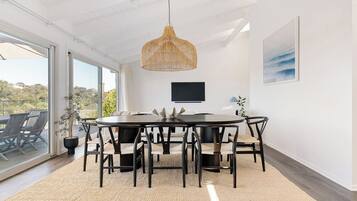 A dining area with a round table, black chairs, a woven pendant light, a flat-screen TV, and a view through large windows.