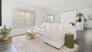 Un salon moderne avec un canapé blanc, une table basse et un tapis. On aperçoit à l’arrière-plan un coin cuisine avec des placards et un plan de travail.