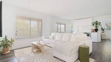 A modern living room with a white sofa, a coffee table, and a rug. There is a kitchen area with cabinets and a countertop visible in the background.
