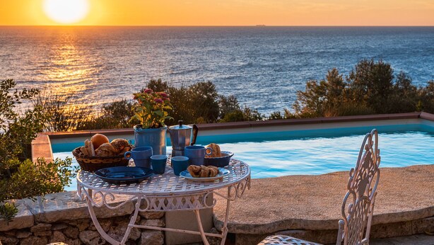 Una mesa dispuesta con alimentos para el desayuno junto a una piscina con vista al atardecer.