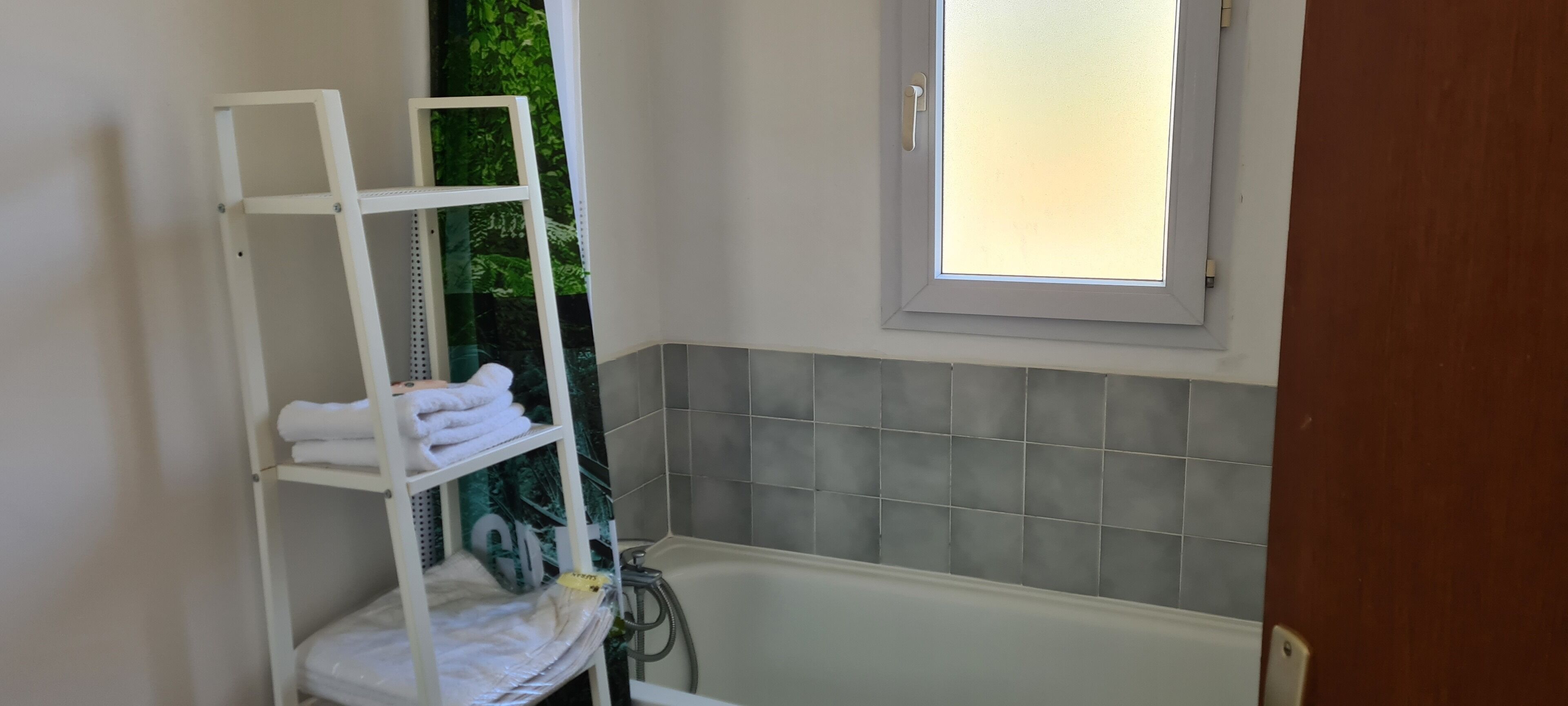 A bathroom with a white shelf holding towels, a bathtub, and a window with frosted glass.