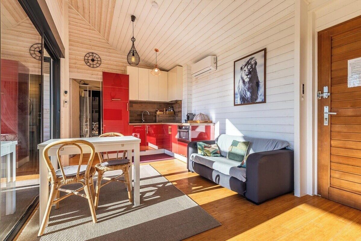 A modern kitchen with red cabinets, a dining area with a table and chairs, and a living room with a sofa and a painting on the wall.