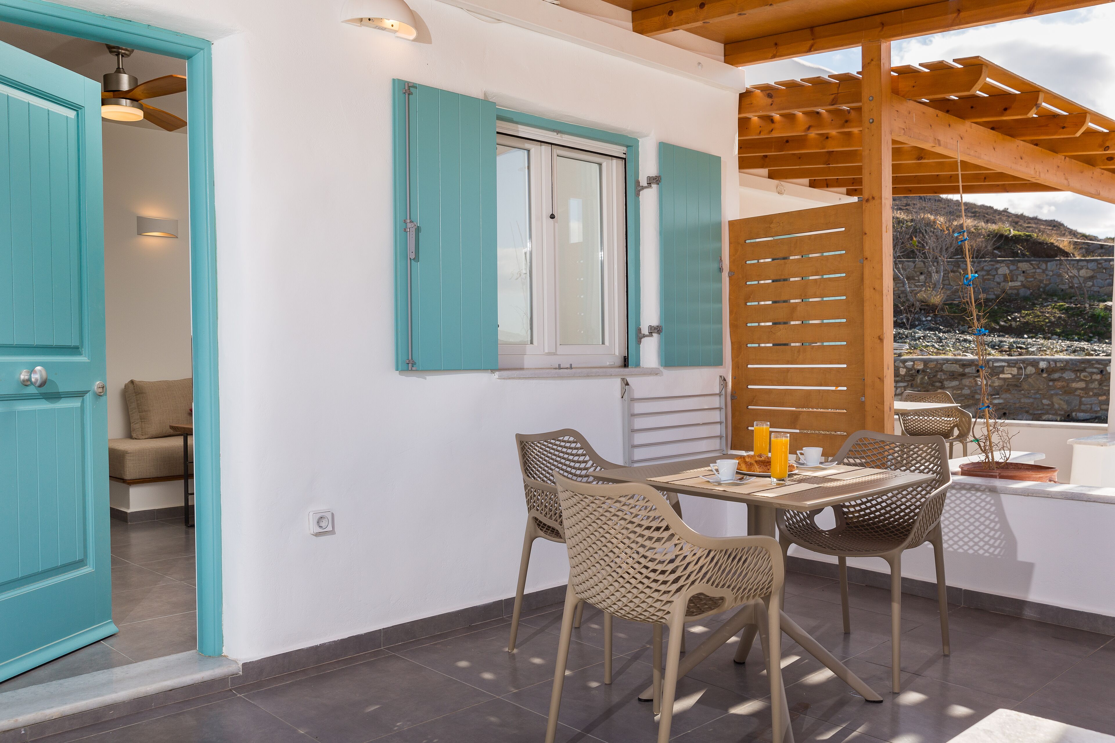A patio with a table and chairs, a window with shutters, and a view of a mountainous landscape.