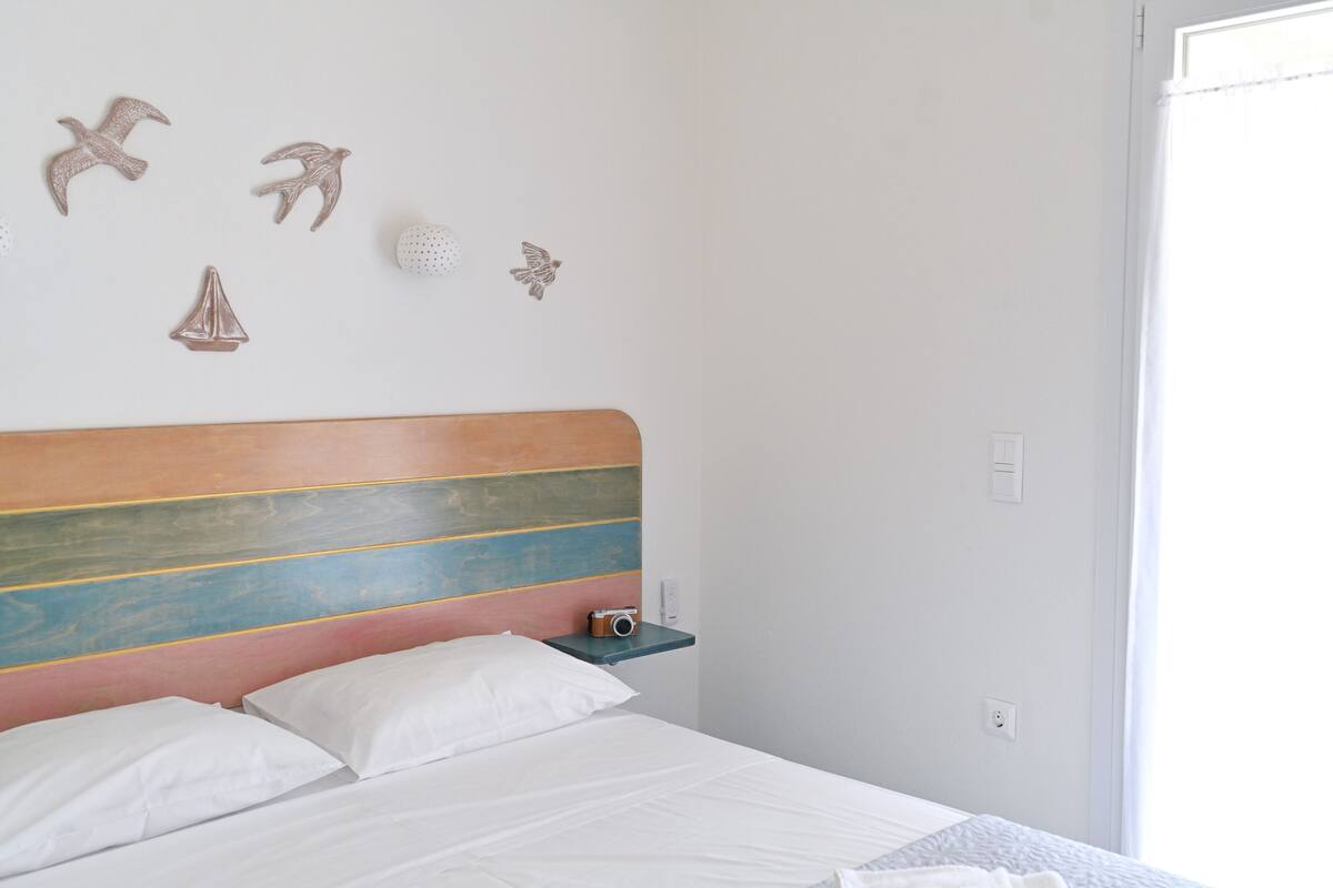 Un lit bien fait, avec du linge de lit blanc, une tête de lit en bois à rayures colorées et une petite table de chevet sur laquelle se trouve un appareil photo.