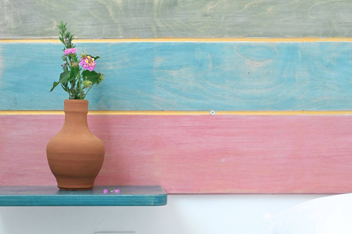 Un vase en terre cuite orné de fleurs roses, posé sur une étagère de couleur turquoise, le tout adossé à un mur à rayures horizontales.