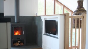 A modern living room with a wood-burning stove, a flat-screen TV mounted on the wall, and a large window providing natural light. - Luxury child and baby friendly rural retreat. Close to Porthleven and St Ives (Helston)