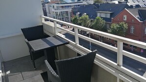 A balcony with two black wicker chairs and a small table, overlooking a street with red brick buildings and a blue sky. - App. Seeblick, Hanseatenhaus (Sylt)