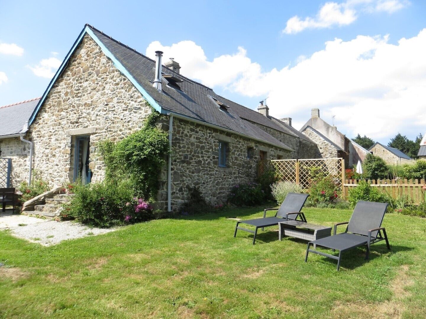 A stone house with a sloped roof and a chimney. There are two lounge chairs in the yard.