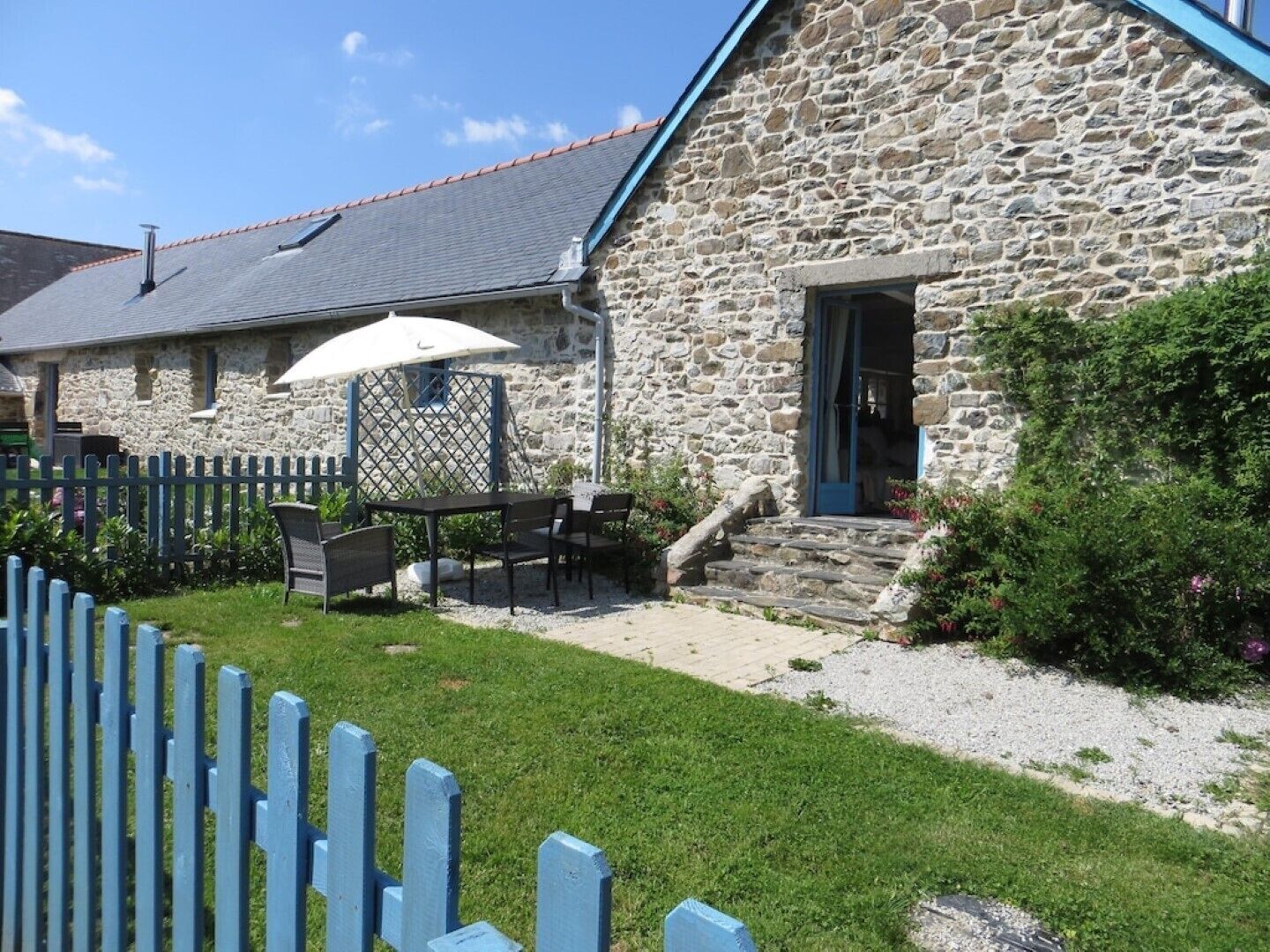 Un bâtiment en pierre avec une porte bleue, un petit espace extérieur aménagé avec une table et des chaises, et un parasol blanc.