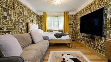 A room with patterned wallpaper, a bed with a headboard, a sofa, a television, and a window with curtains.