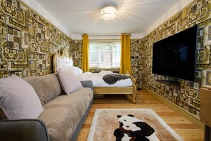 Een kamer met behang met patroon, een bed met hoofdeinde, een bank, een televisie en een raam met gordijnen.