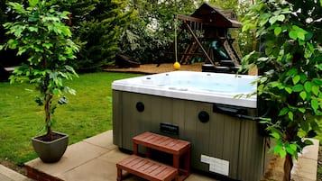 A hot tub with a wooden deck and a small bench, surrounded by lush greenery and a wooden play structure in the background.