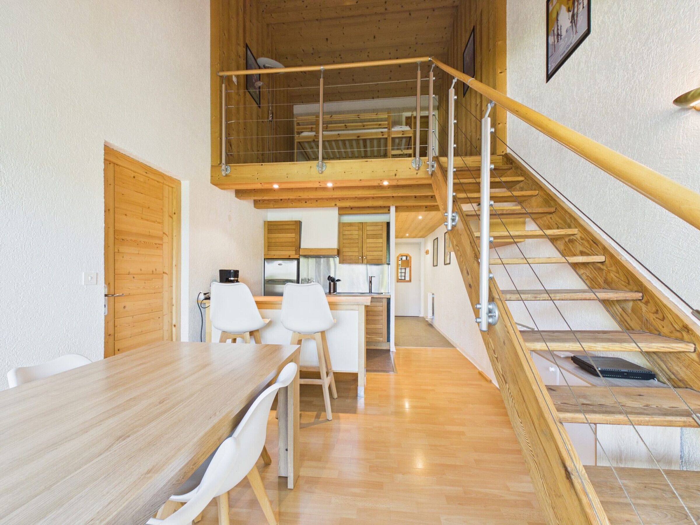 Un interno moderno con un tavolo da pranzo in legno, sedie bianche, una zona cucina e una scala in legno.