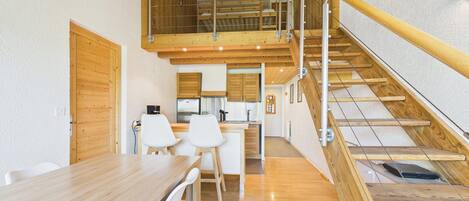 Un interno moderno con un tavolo da pranzo in legno, sedie bianche, una zona cucina e una scala in legno.