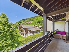 Un balcon donnant sur un village perché sur une colline, avec des balustrades en bois et un petit espace extérieur aménagé pour s’asseoir.