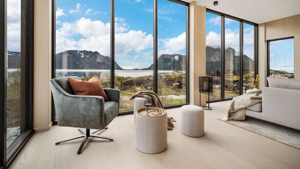A modern living room with large windows, a grey armchair, a round ottoman, and a beige sofa. - NEW! Luxury Cabin in beautiful Lofoten (Nordland)
