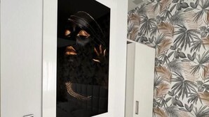 Una habitación con pared blanca, una obra de arte enmarco, una mesa blanca con lámpara, un mueble blanco y una pared con diseño de hojas tropicales.