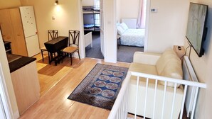 Un ambiente compatto con angolo cottura, zona pranzo, camera da letto con un letto e un comodino, e soggiorno con divano bianco e televisione.