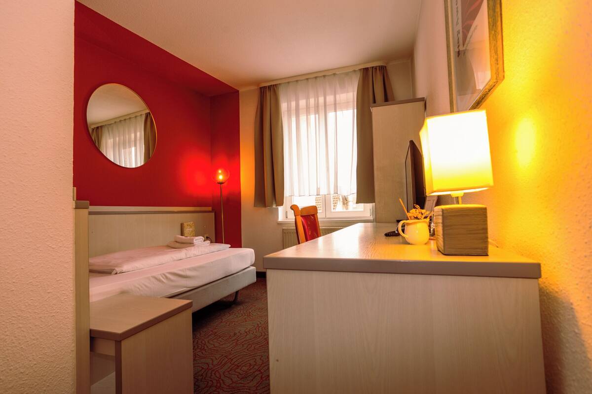 A hotel room with a bed, a desk, a chair, a mirror, a window with curtains, and a lamp.