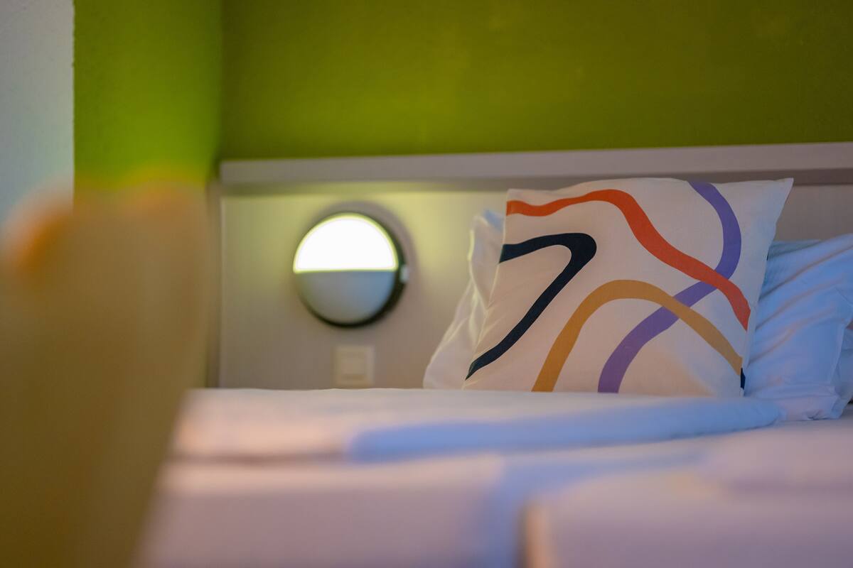 A bed with a white headboard, a round mirror, and a pillow with a multicolored design.