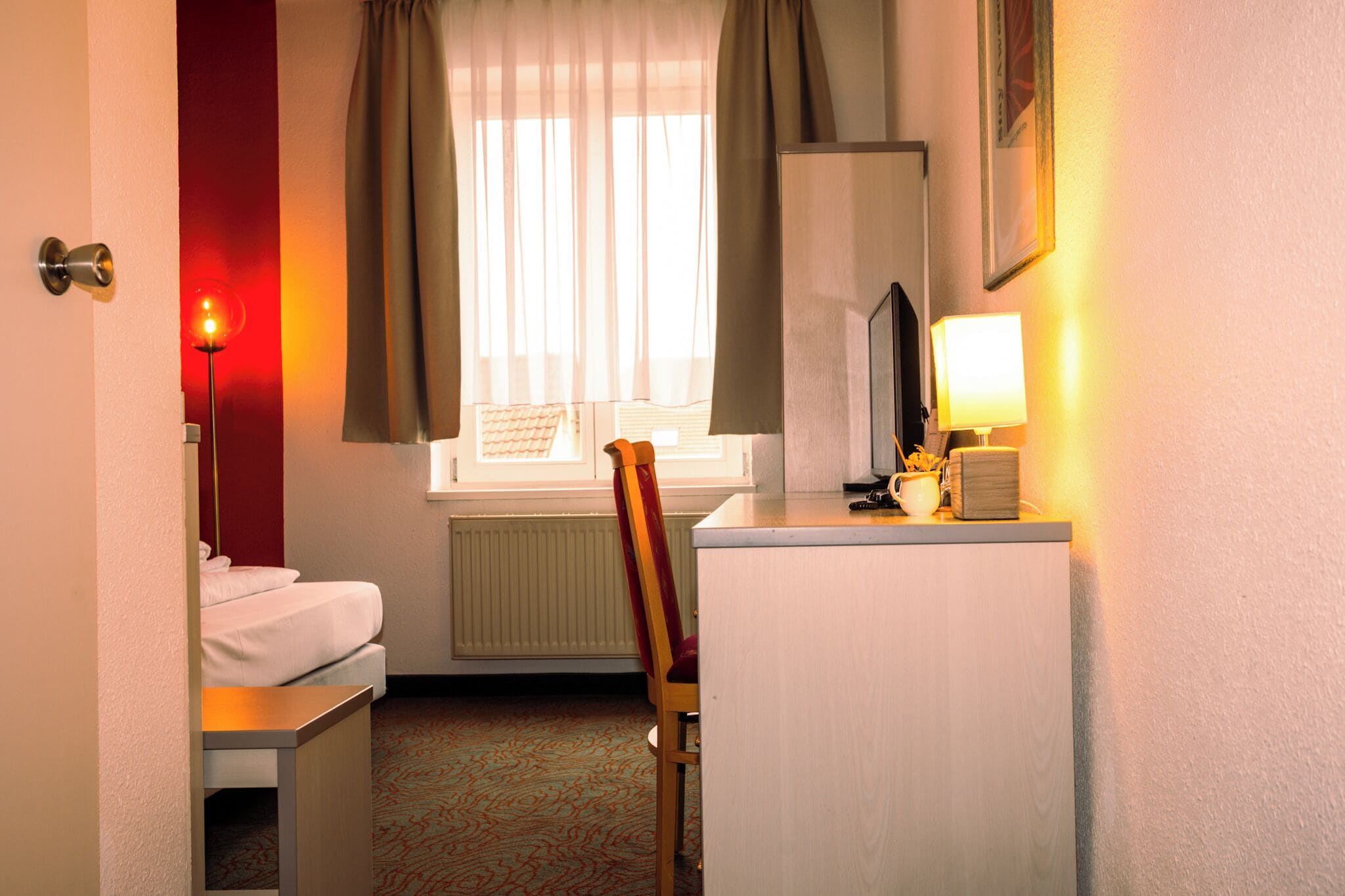 Une chambre d’hôtel avec un lit, un bureau avec une lampe, une chaise et une fenêtre avec des rideaux.