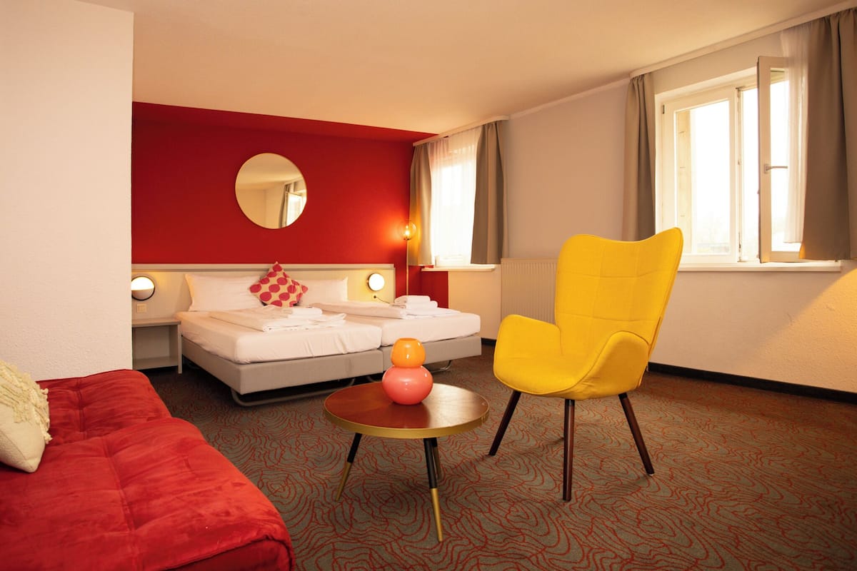 A hotel room with a red wall, a round mirror, a bed with white bedding, a yellow armchair, and a small round table with a pink vase.