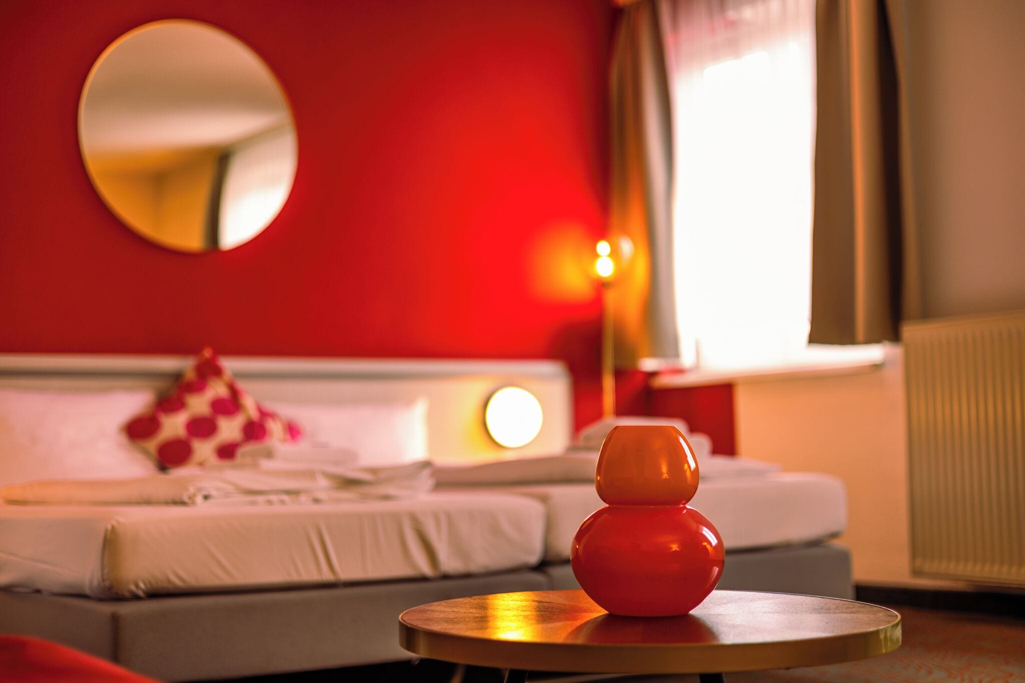 A hotel room with a red wall, a round mirror, a bed with white bedding, and a small table with a red vase.