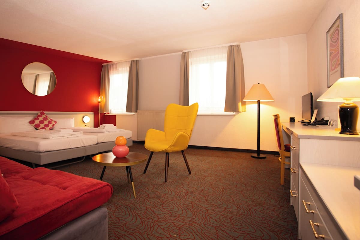 A hotel room with a bed, a yellow chair, a desk with a lamp, and a red sofa.