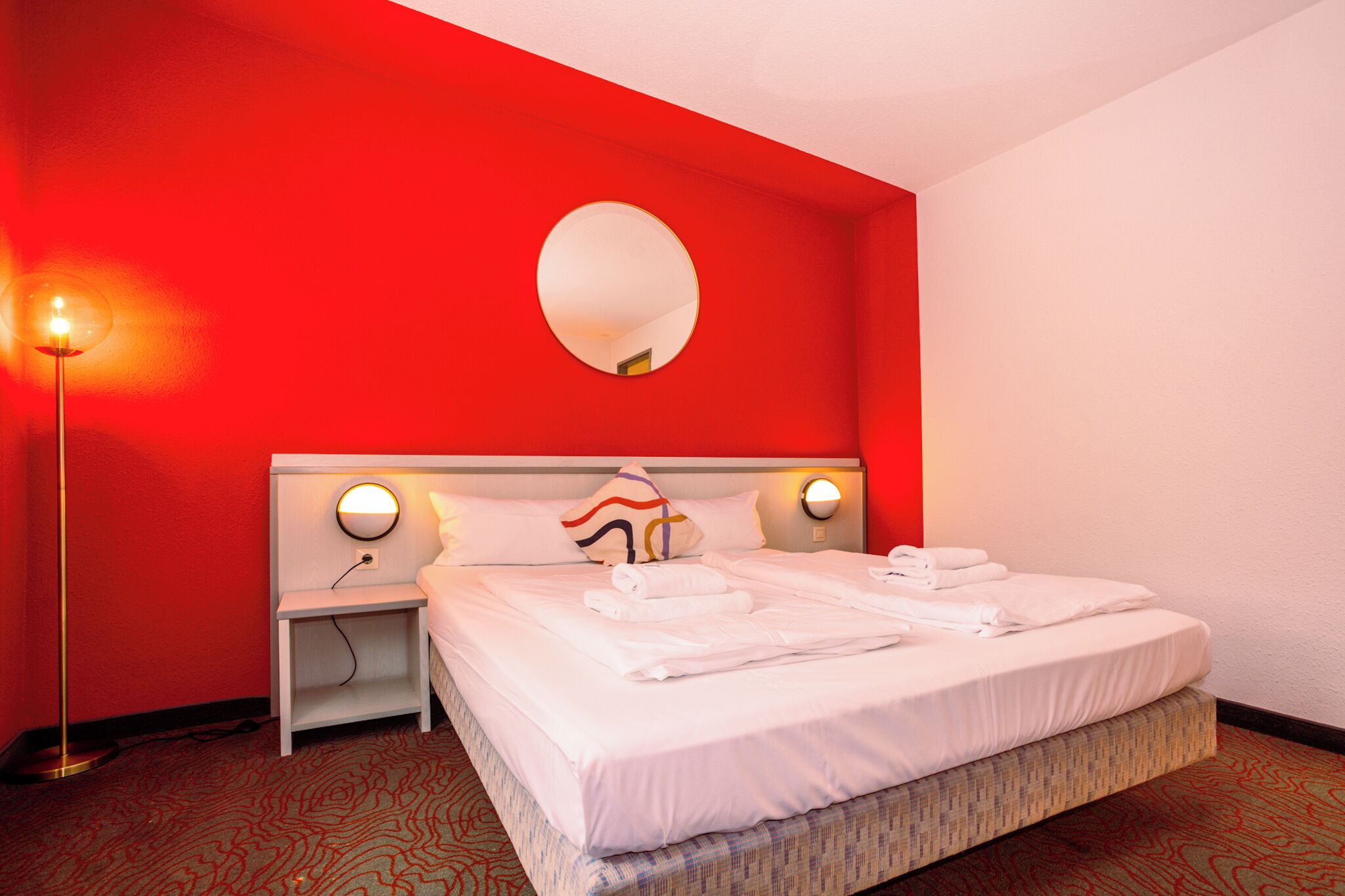Habitación de hotel con una pared de acento roja, un espejo redondo, una cama con toallas blancas ordenadas y una lámpara de pie.