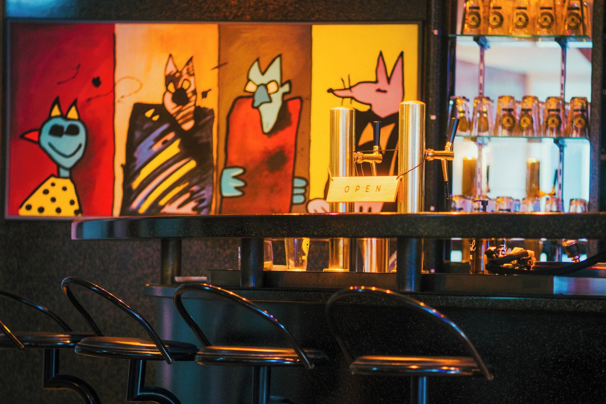 Una zona de bar con mostrador, taburetes y una estantería llena de botellas.