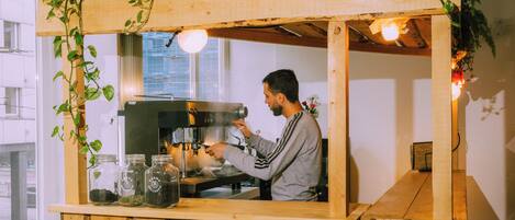 Une personne utilise une machine à café derrière un comptoir en bois, devant lequel se trouvent des bocaux remplis de grains de café et une plante suspendue.