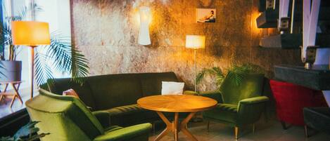 Eine Lobby mit einem grünen Sofa, zwei Sesseln, einem Holztisch und einer Marmorwand mit Wandleuchten.
